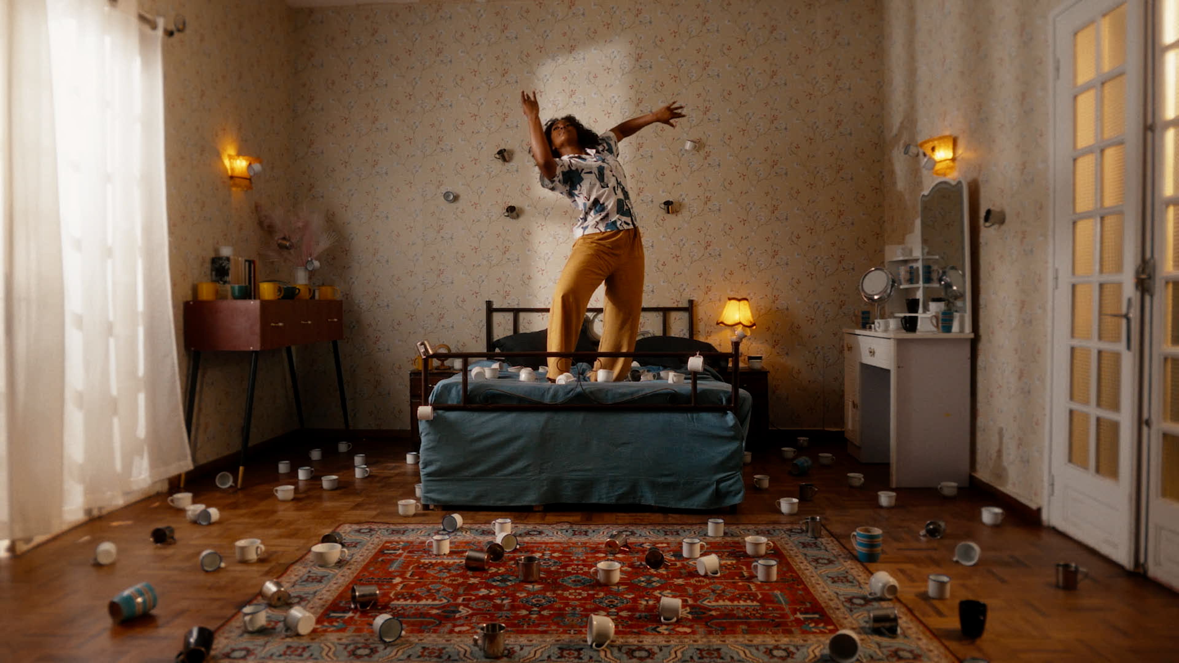 Une femme danse sur un lit dans une chambre dont le sol est couvert de tasses